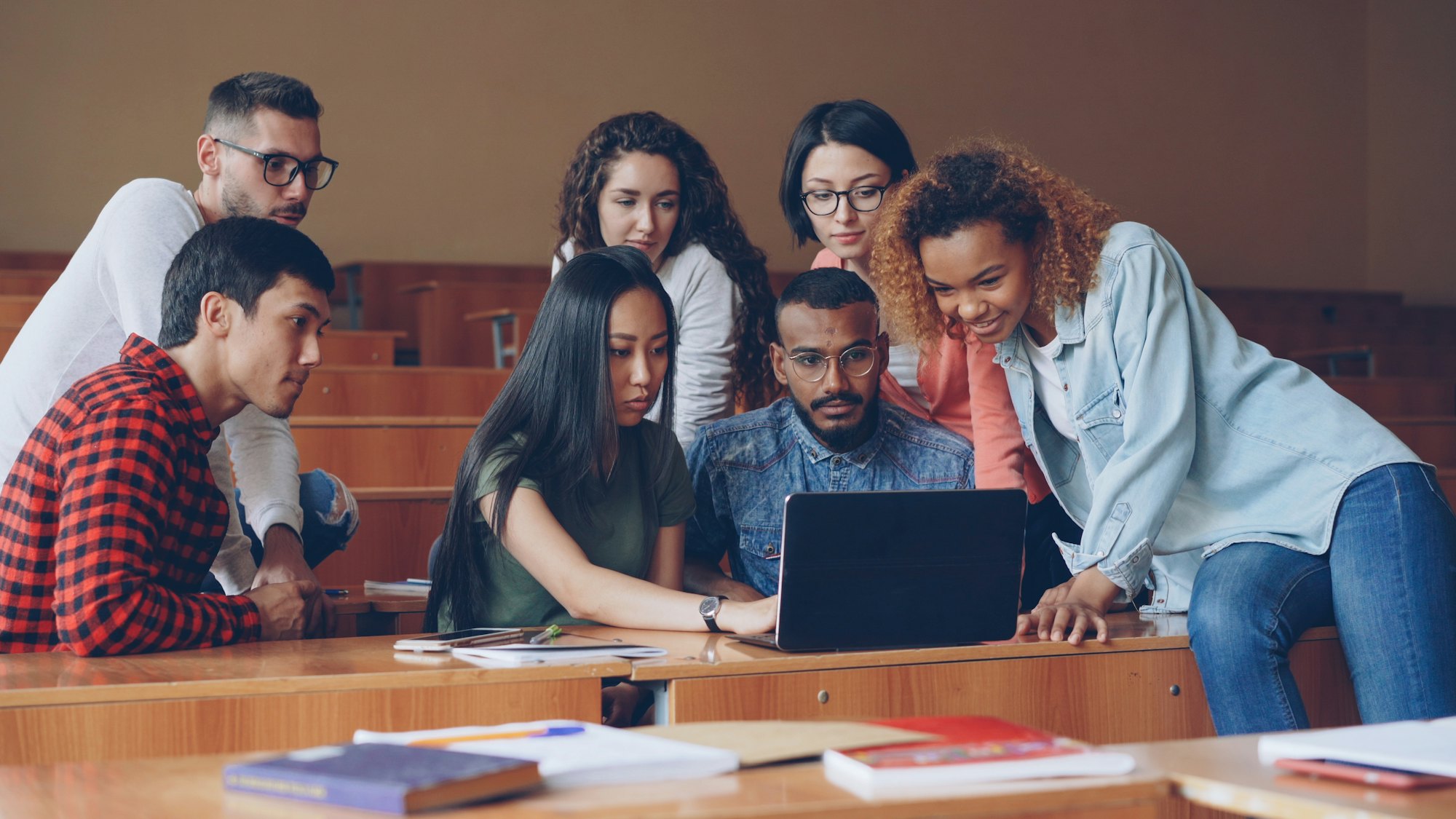 A group of counselling students gathered around a laptop together in a warm, sunlit lecture room.